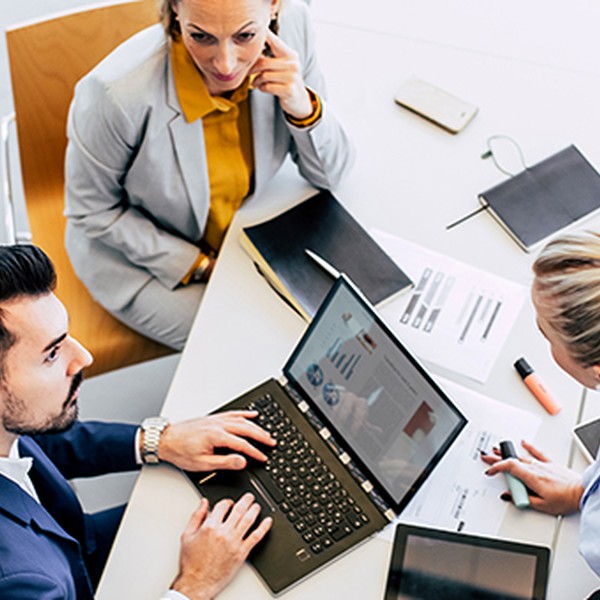 A laptop displays graphs as three professionals collaborate around a table, one typing, while the others discuss. The setting is modern, with various stationery items and a large window in view.
