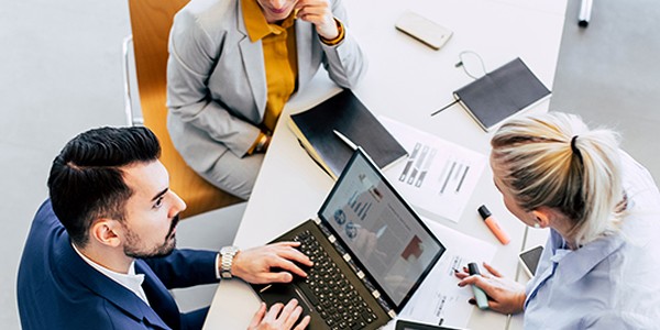 A laptop displays graphs as three professionals collaborate around a table, one typing, while the others discuss. The setting is modern, with various stationery items and a large window in view.