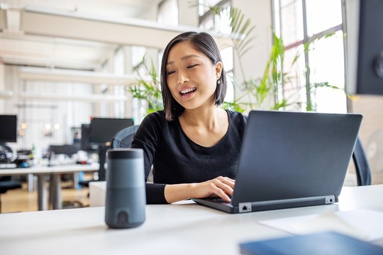 A woman with short black hair smiles while typing on a laptop. A smart speaker sits on the table in a bright, modern office filled with plants and desks.