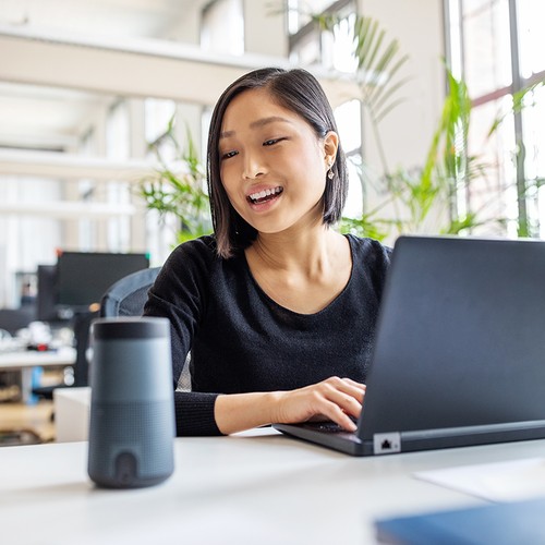 A woman with short black hair smiles while typing on a laptop. A smart speaker sits on the table in a bright, modern office filled with plants and desks.