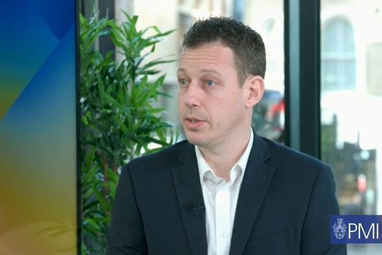 A man in a black suit is speaking, seated in front of a window with a blurred urban background. Green plants are visible nearby, and a logo for "PMI tv" is in the corner.