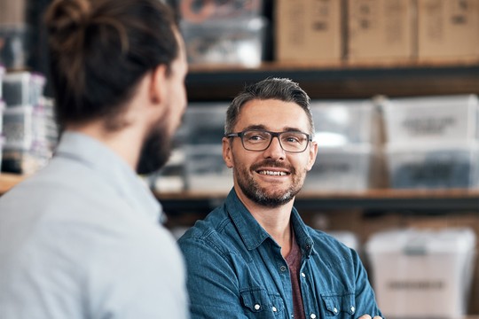 A man with glasses and a beard smiles while engaging in conversation with another man. They are indoors, surrounded by boxes and storage containers in a workshop-like environment.