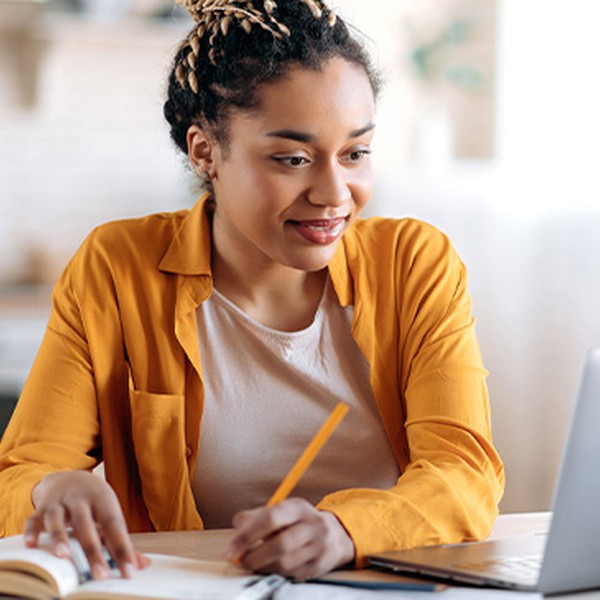 A woman in a yellow shirt sits at a table, writing in a notebook while looking at a laptop. She is in a bright, modern kitchen with soft natural light.