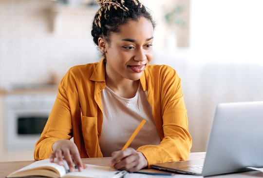 A woman in a yellow shirt sits at a table, writing in a notebook while looking at a laptop. She is in a bright, modern kitchen with soft natural light.