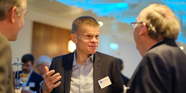A man in a suit engages in conversation, gesturing with his hands. He is surrounded by other men in a well-lit indoor setting, likely at a formal gathering or event.