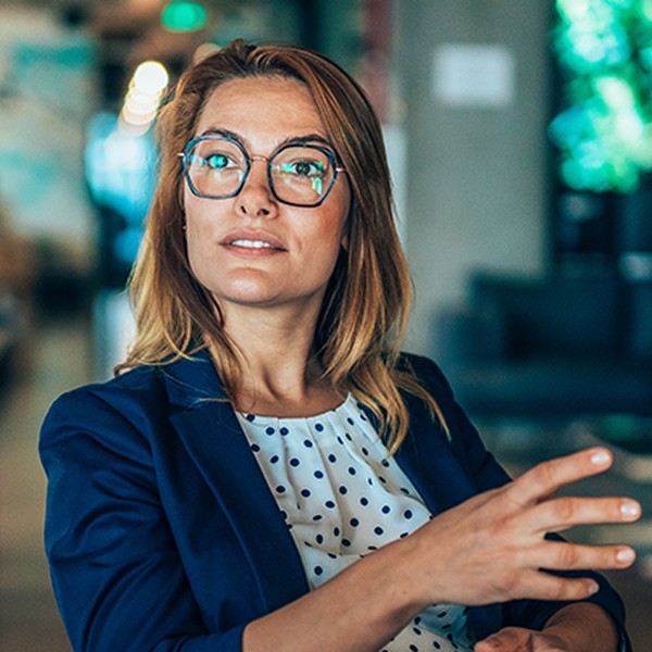 A woman with blonde hair and glasses gestures while speaking in a modern indoor setting. She wears a blazer over a polka-dot blouse. Greenery and soft seating are visible in the background.