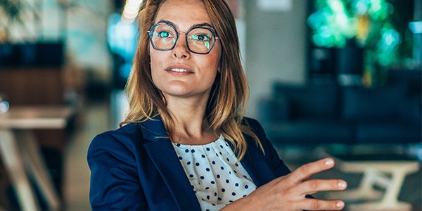 A woman with blonde hair and glasses gestures while speaking in a modern indoor setting. She wears a blazer over a polka-dot blouse. Greenery and soft seating are visible in the background.
