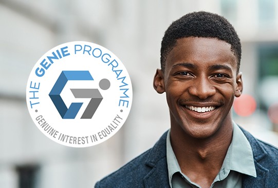 A young man with a broad smile stands in an urban environment. He is wearing a jacket over a shirt. The logo of "The Genie Programme" is displayed prominently beside him.