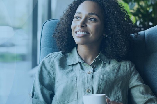 A woman sits comfortably in a chair, holding a cup while smiling. She is in a bright space with large windows, greenery visible outside, conveying a relaxed, contemplative atmosphere.