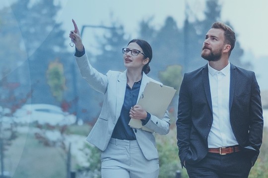 A woman in a suit gestures while talking, holding a folder. A man in a suit walks beside her. They are outdoors in a professional setting with modern buildings and greenery.