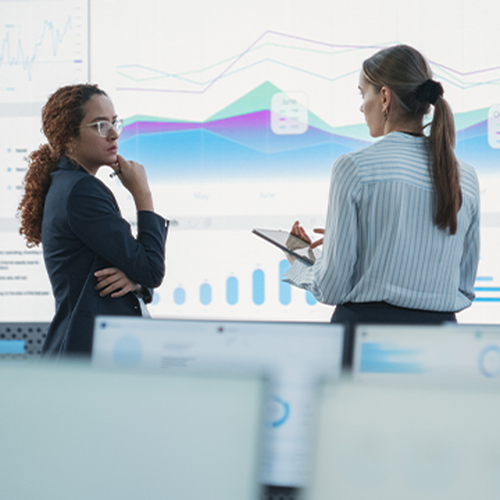 Two professionals discuss data trends displayed on large digital screens in an office. One woman stands with arms crossed, while the other holds a tablet and gestures toward the graphs.