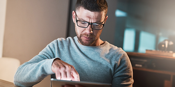 A man wearing glasses is focused on a tablet, using his fingers to interact with the screen. He is seated in a well-lit indoor space, with modern furnishings visible in the background.