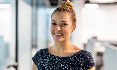 A woman with a bun hairstyle smiles warmly while wearing a polka-dotted top. She stands in a modern office space with blurred desks and soft lighting in the background.