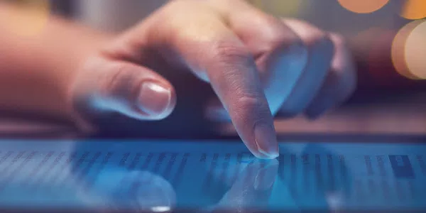 A finger gently touches a glowing tablet screen displaying text. The setting is softly lit, suggesting a cozy atmosphere with blurred background lights.