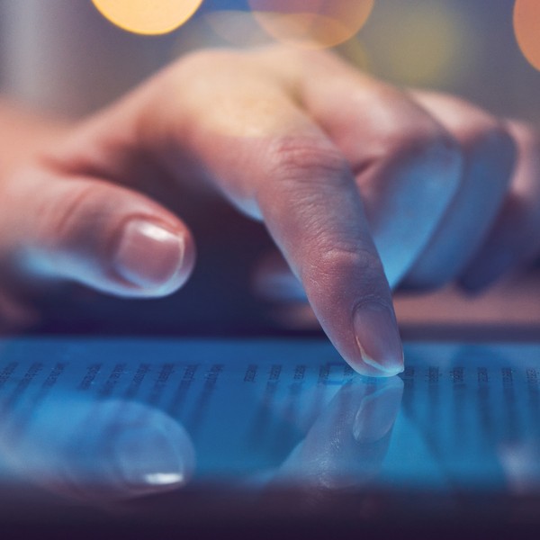 A finger gently touches a glowing tablet screen displaying text. The setting is softly lit, suggesting a cozy atmosphere with blurred background lights.