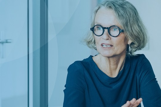 A middle-aged woman with glasses gestures while speaking in a modern conference room, with a whiteboard and light blue walls creating a professional atmosphere.
