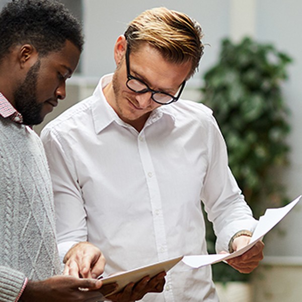 Two men are examining documents together. One man, wearing a gray sweater, points at the papers while the other, in a white shirt, closely inspects them. They are in a modern, well-lit indoor space with greenery in the background.
