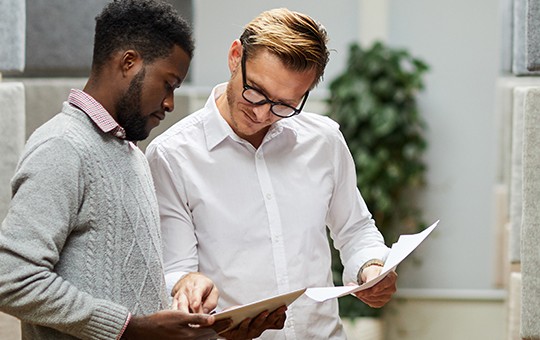 Two men are examining documents together. One man, wearing a gray sweater, points at the papers while the other, in a white shirt, closely inspects them. They are in a modern, well-lit indoor space with greenery in the background.
