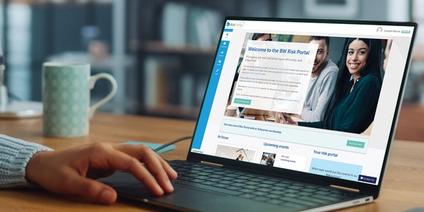 A hand is typing on a laptop displaying the BW Risk Portal webpage. In the background, there’s a patterned mug and blurred shelves, suggesting a cozy workspace.