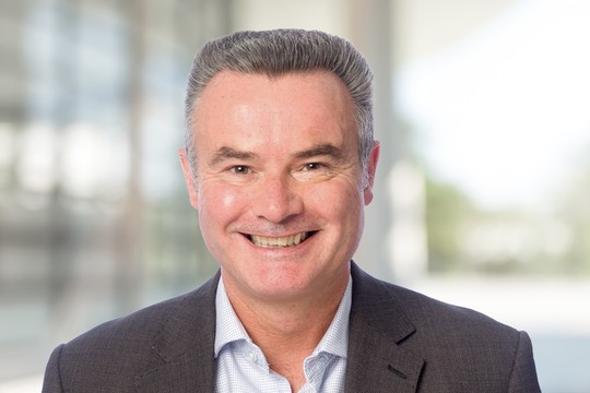 A middle-aged man smiles broadly, wearing a dark blazer over a light shirt. He stands against a blurred background suggesting a modern indoor setting.