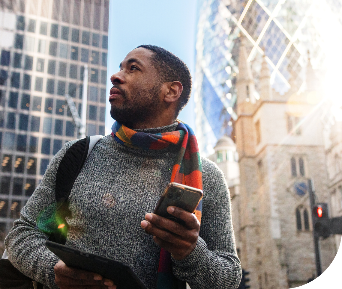 A man stands in a busy urban environment, looking upward while holding a smartphone and a tablet. Modern glass buildings and a historical structure are in the background.