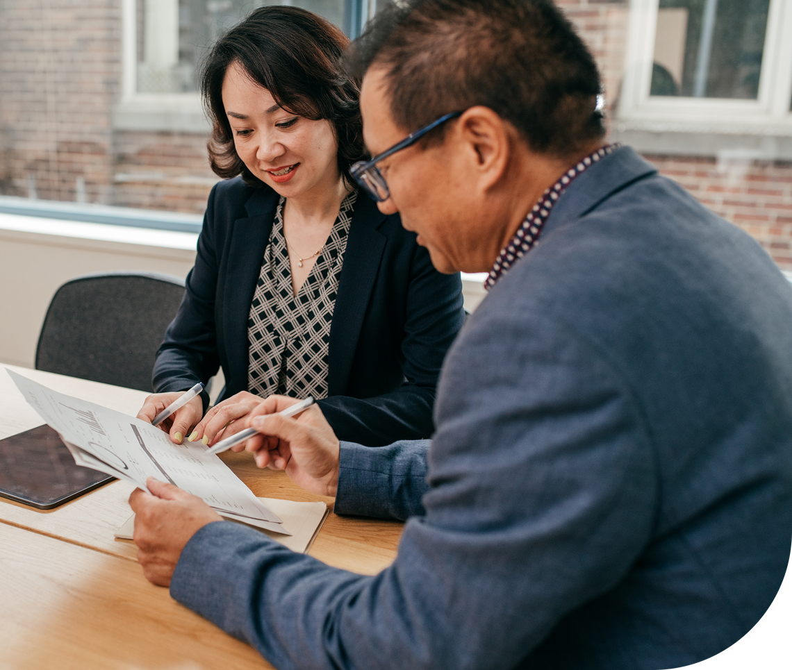 A woman and a man sit at a wooden table, reviewing documents together. Natural light filters in through a window, illuminating their focused expressions and the papers they are discussing.