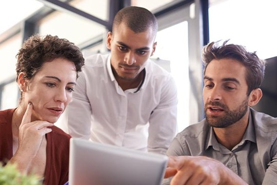 A group of three people, one woman and two men, are attentively looking at a tablet. They are engaged in discussion within a modern, bright office environment.