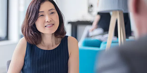 A woman with shoulder-length hair smiles while holding a tablet, engaged in conversation at a modern café. Bright light streams through large windows, illuminating stylish furniture in the background.