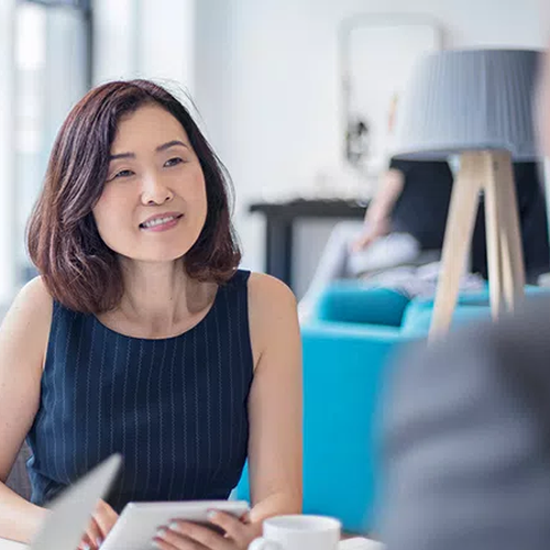 A woman with shoulder-length hair smiles while holding a tablet, engaged in conversation at a modern café. Bright light streams through large windows, illuminating stylish furniture in the background.