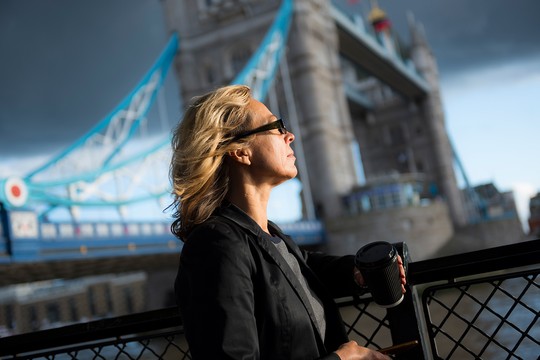 A woman holds a coffee cup while gazing thoughtfully towards the Tower Bridge in a dynamic setting, with a stormy sky creating a dramatic backdrop.