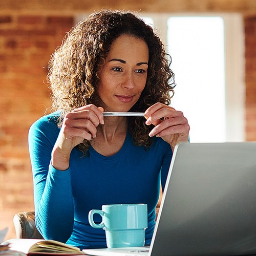 A woman sits at a desk, holding a pen and looking thoughtfully at a laptop screen. A blue mug and some open books are nearby, surrounded by a cozy, brick-walled environment.