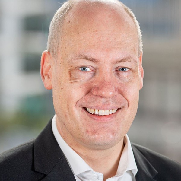 A smiling man with short, light-colored hair and blue eyes wears a black suit jacket over a white shirt. He is positioned in an indoor environment with a blurred, modern background.