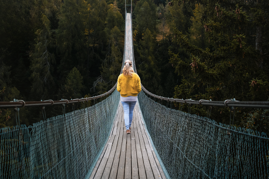 A woman in a yellow sweater walks along a long suspension bridge made of wood and netting, surrounded by dense, green trees and forest scenery.