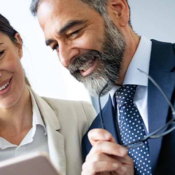 A man and woman are smiling while looking at a tablet, suggesting collaboration. A blurred individual in business attire is positioned behind them, indicating a professional office setting with natural light.