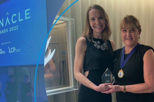 Two women stand together, one holding an award, in a decorated banquet hall. The backdrop features a large banner reading “PINNACLE Awards 2022,” indicating a formal event.