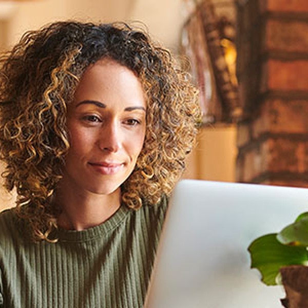 A woman with curly hair focuses intently on a laptop while seated in a cozy, well-lit room. A small green plant sits beside her, enhancing the homey atmosphere.