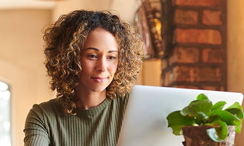 A woman with curly hair focuses intently on a laptop while seated in a cozy, well-lit room. A small green plant sits beside her, enhancing the homey atmosphere.