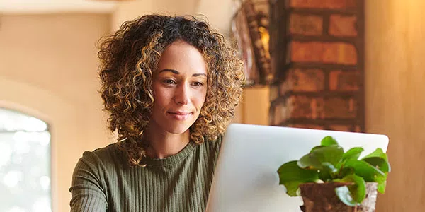 A woman with curly hair focuses intently on a laptop while seated in a cozy, well-lit room. A small green plant sits beside her, enhancing the homey atmosphere.