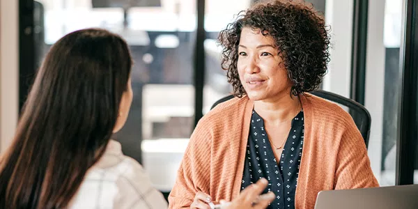 Two women are engaged in a conversation in a modern office setting. One woman, wearing a brown cardigan, appears to be actively listening and responding to the other, who sits across from her.