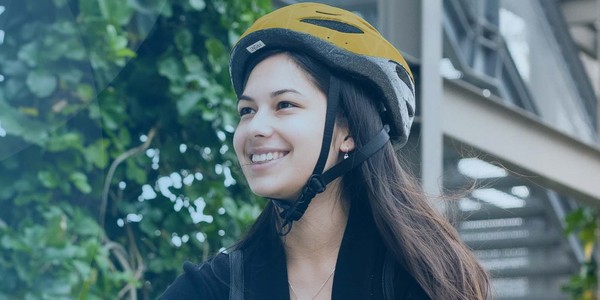 A young woman wearing a yellow bicycle helmet smiles while standing outdoors. She is surrounded by greenery, and there are structures in the background, suggesting a relaxed, active environment.