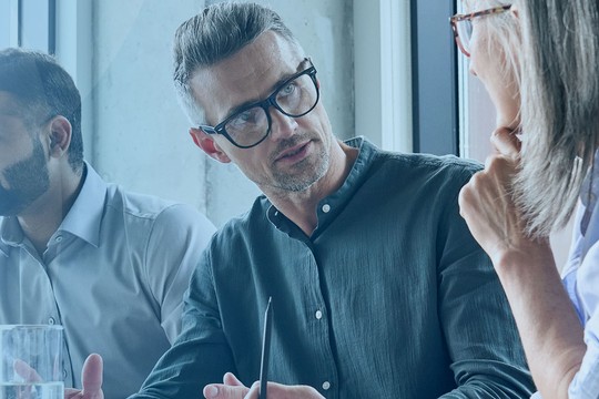 Three professionals are engaged in a discussion around a conference table, with a glass of water in the foreground and large windows providing natural light in a modern office environment.