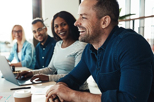 A group of four people sits at a table, engaged in conversation and smiling. Laptops and a coffee cup are nearby. Bright natural light enters from large windows, creating a welcoming atmosphere.