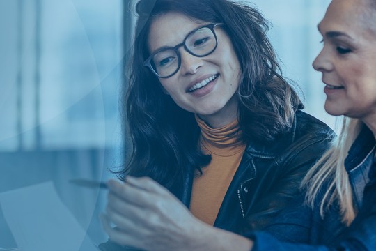 Two women are engaged in a discussion, one smiling and pointing at an object. They are seated at a table in a well-lit, modern environment with large windows.