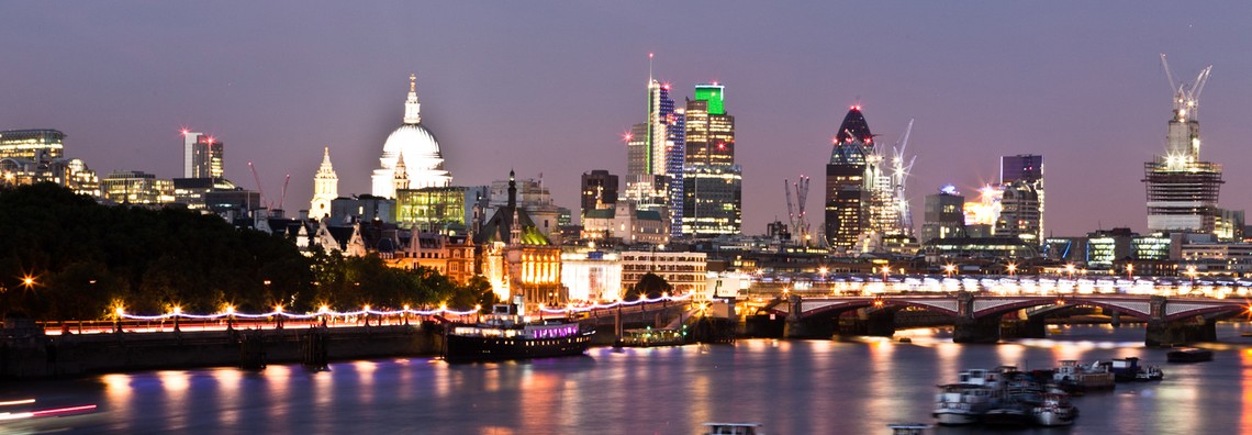 A city skyline features illuminated buildings and a prominent dome, with reflections on the water. Boats are seen on the river, and a bridge connects both shores at dusk.