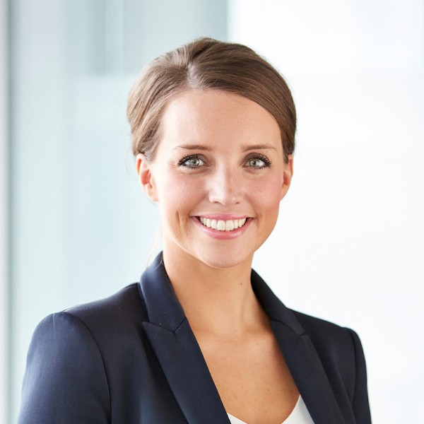 A smiling woman with brown hair, dressed in a dark blazer over a white top, stands against a softly-lit modern interior, conveying professionalism and approachability.