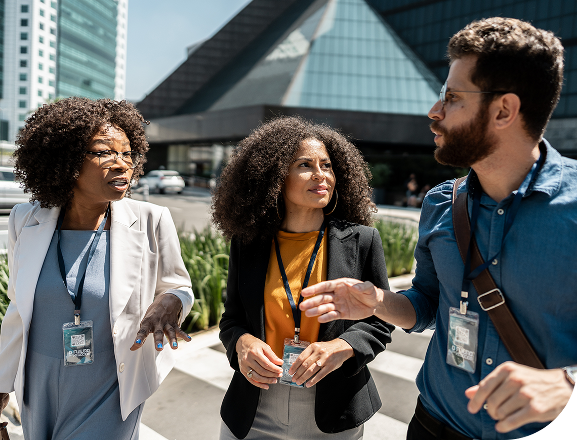Three professionals engage in conversation while walking on a city sidewalk. They appear to discuss something animatedly, with modern buildings and greenery in the background under a clear sky.