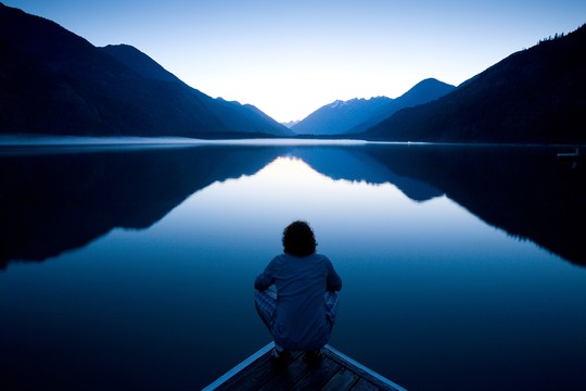 A person sits quietly on a dock, gazing at a serene lake reflecting surrounding mountains and a twilight sky, creating a peaceful and contemplative atmosphere.