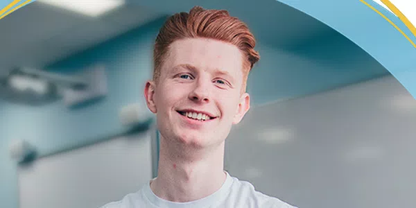 A young man with light red hair smiles warmly, standing against a softly lit classroom background with whiteboards visible. His casual white shirt suggests a relaxed, friendly atmosphere.
