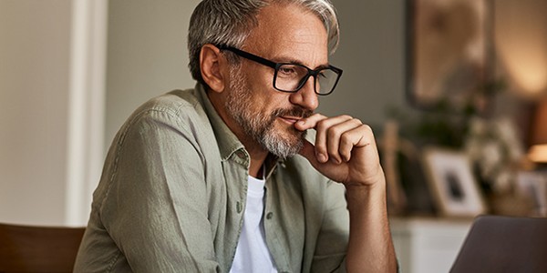 A middle-aged man with glasses sits at a table, thoughtfully resting his chin on his hand while gazing at a laptop. Soft, warm lighting and home decor create a cozy atmosphere.