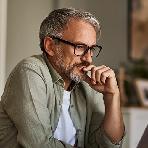 A middle-aged man with glasses sits at a table, thoughtfully resting his chin on his hand while gazing at a laptop. Soft, warm lighting and home decor create a cozy atmosphere.